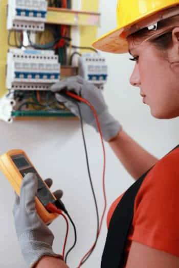 Electrician in a yellow hard hat using a multimeter to test a circuit board for electrical issues.