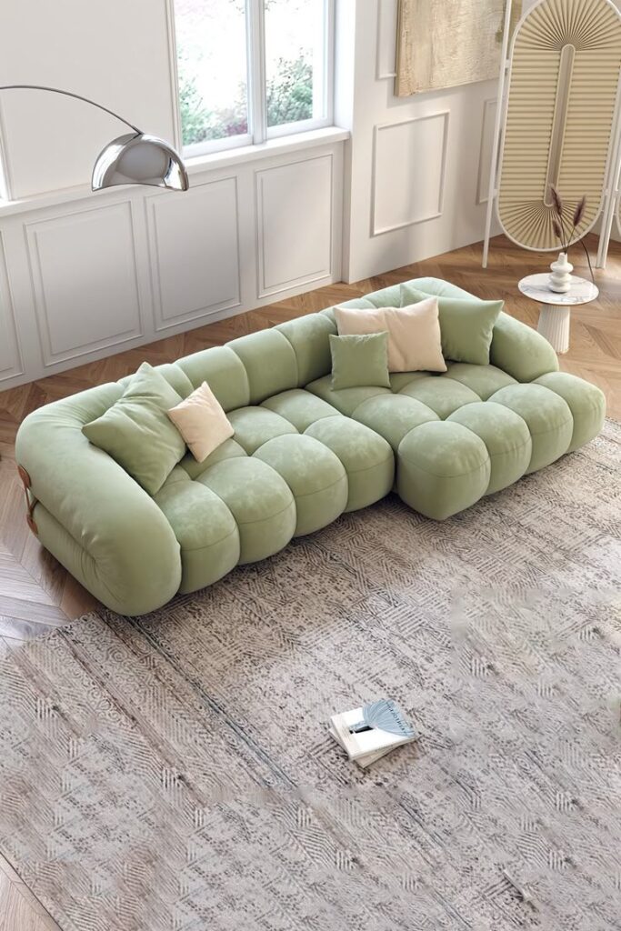 Modern living room with green tufted sofa, beige pillows, textured rug, and natural light from large window.