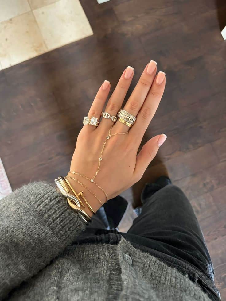 Hand with elegant gold and silver rings and bracelets, featuring polished nude nails and a cozy gray sweater background.