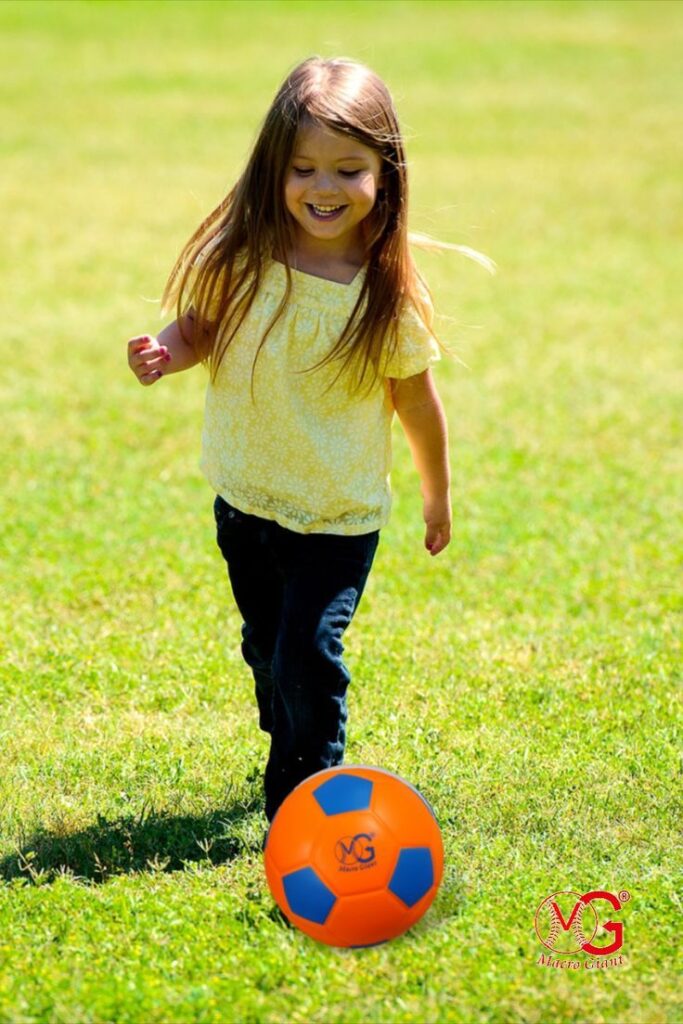 Young girl playing soccer on a grassy field, smiling and chasing an orange and blue ball.