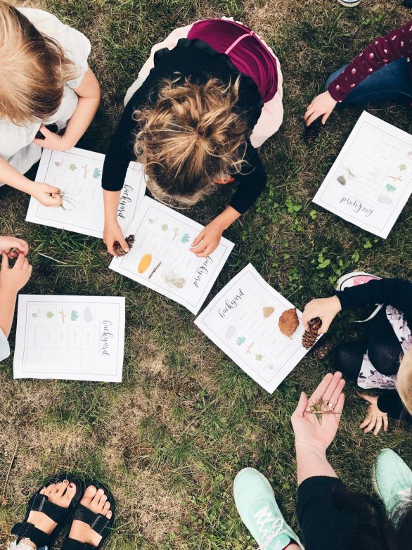 Children engaging in outdoor scavenger hunt with worksheets and natural items on a grassy field.