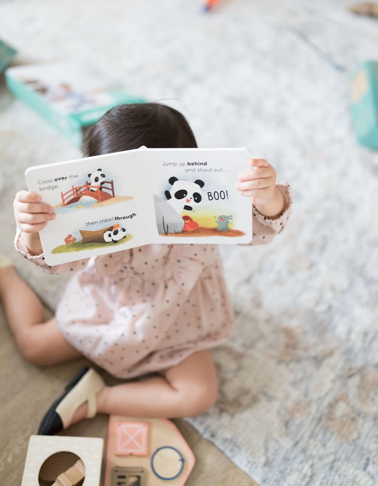 Child reading a picture book with panda illustrations, surrounded by toys on a carpeted floor.