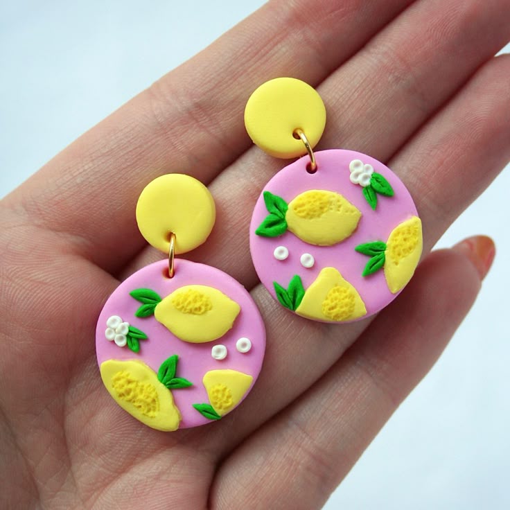 Colorful lemon-themed polymer clay earrings on fingers, featuring pink, yellow, and green designs.