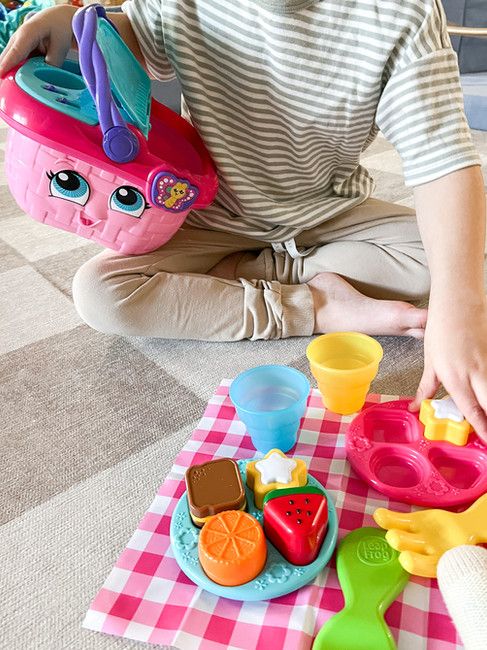 Child playing with a colorful toy picnic set on a checkered blanket, featuring cups and food shapes.