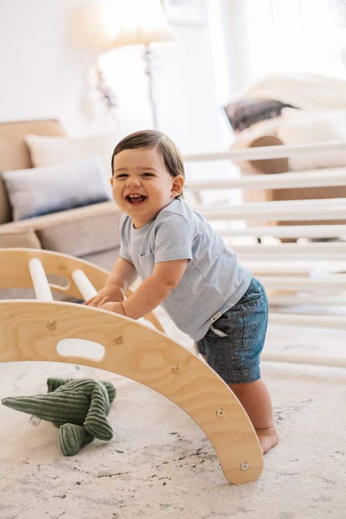 Cheerful toddler playing on wooden toy arch indoors, near plush alligator.