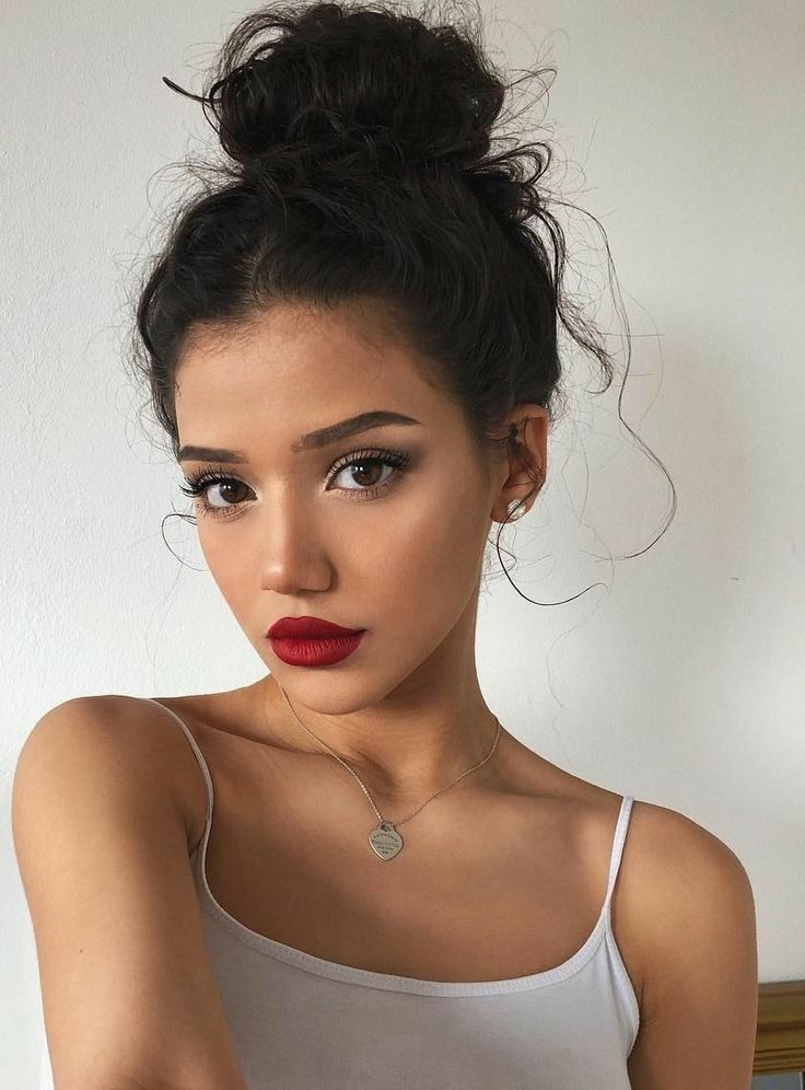 Smiling woman with red lipstick and bun hairstyle wearing a white top, posing against a plain background.