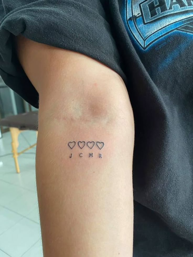 Tattoo of four hearts and the letters JCMR on a person's arm, wearing a dark shirt.