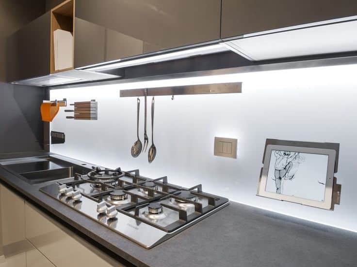 Modern kitchen with gas stove, utensils hanging, and tablet stand on sleek countertop with under-cabinet lighting.