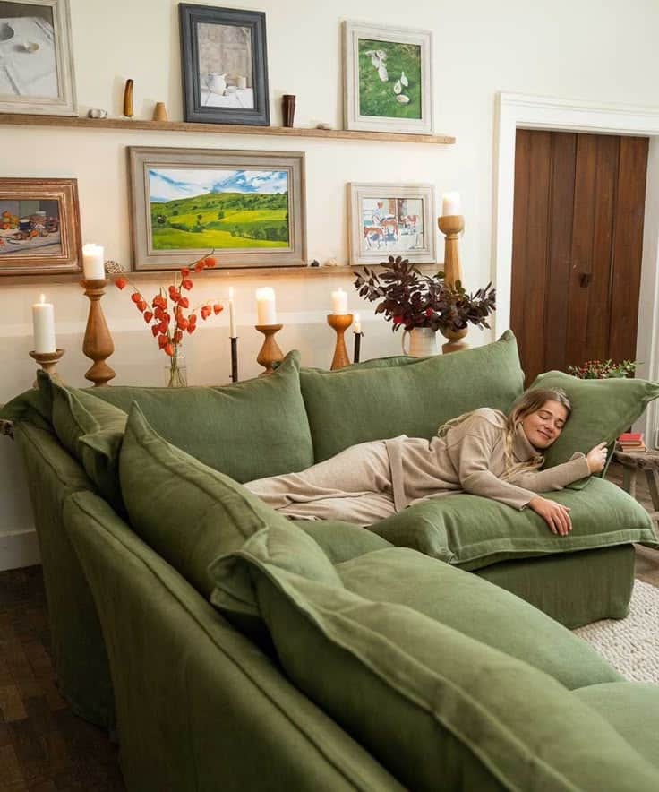 Woman relaxing on a cozy green couch in a stylish, art-filled living room with warm lighting and decor.