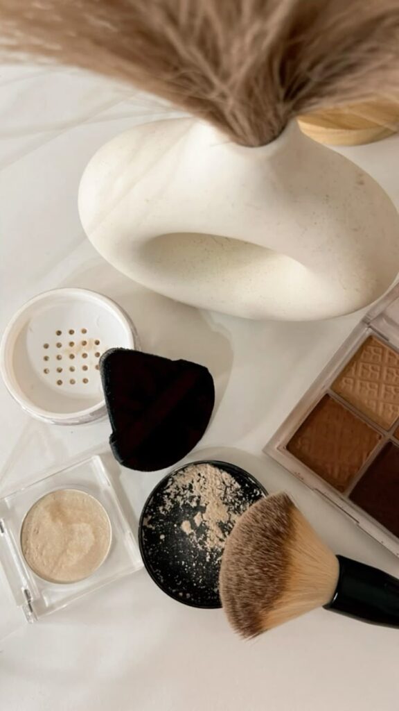 Makeup essentials: loose powder, compact, contour palette, and brush on a white surface with a vase.