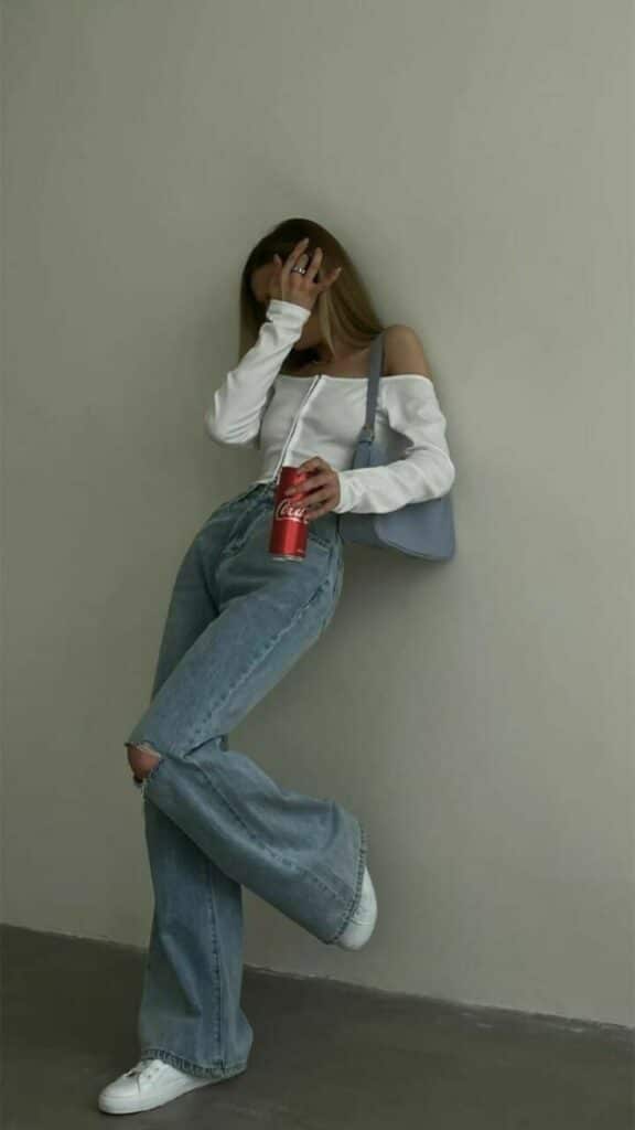 Stylish woman in white top and jeans with a red soda can, leaning against a wall for a casual, trendy look.