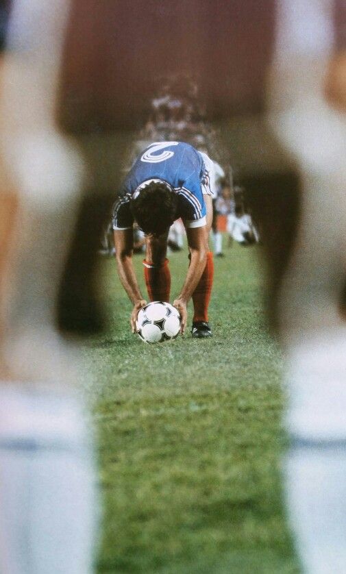 Soccer player prepares to kick ball on field during match, captured through legs of teammates, wearing blue jersey.