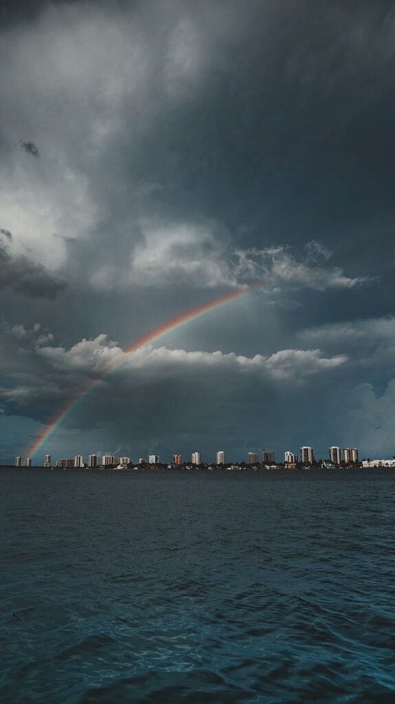 Rainbow arcs over coastal city skyline beneath dramatic cloudy sky, reflecting in calm ocean water.