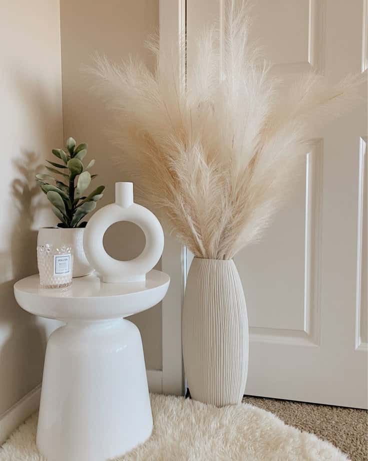Minimalist decor with white vase, pampas grass, candle, and plant on white table against a neutral background.
