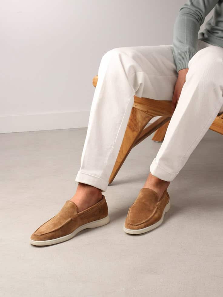 Man in white pants sitting, showcasing stylish brown suede loafers on a modern wooden bench.