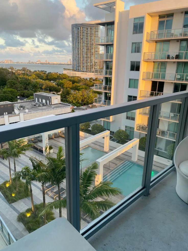Modern balcony view of a pool, city buildings, and lush greenery under a bright, cloudy sky.