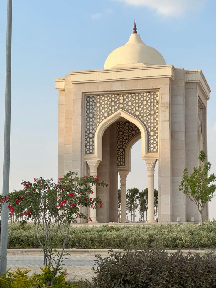 Elegant stone pavilion with intricate arches and dome surrounded by greenery under a clear blue sky.