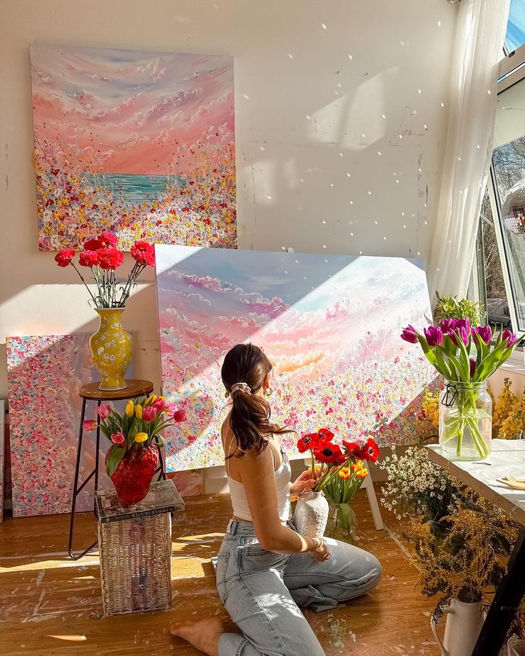Artist arranging flowers in a bright studio, surrounded by vibrant landscape paintings and sunlight.