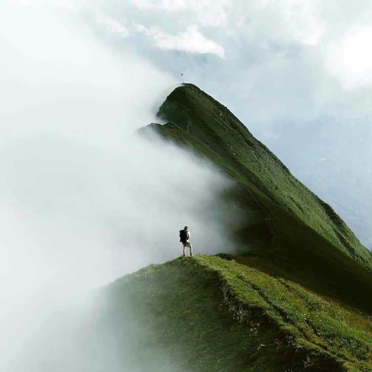 Hiker standing on a misty green mountain ridge, enveloped by clouds, under a cloudy sky.