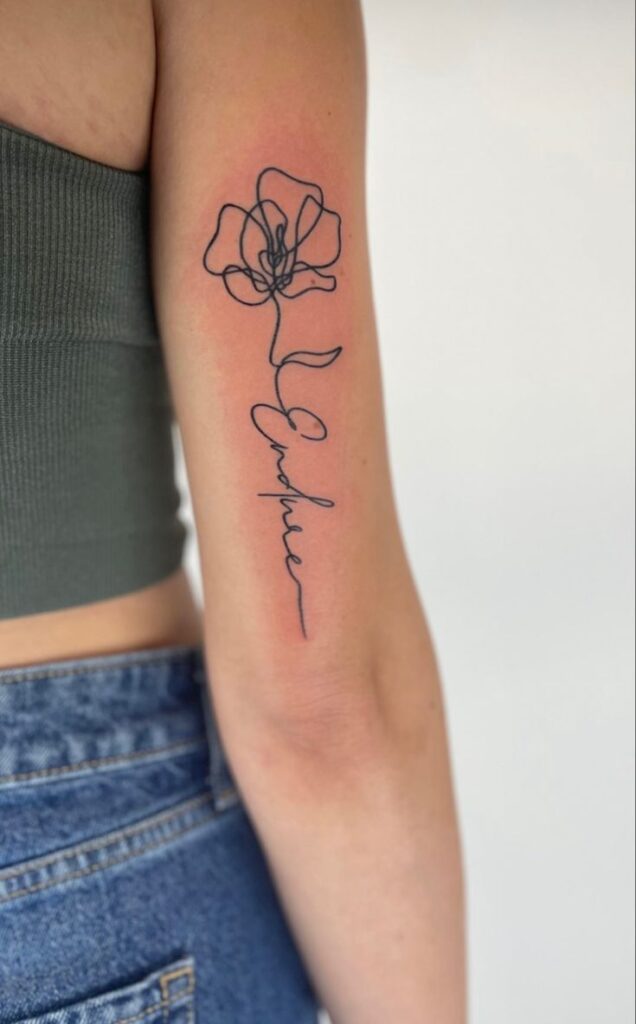 Minimalist flower and Embrace tattoo on woman's arm, wearing a green top and jeans.