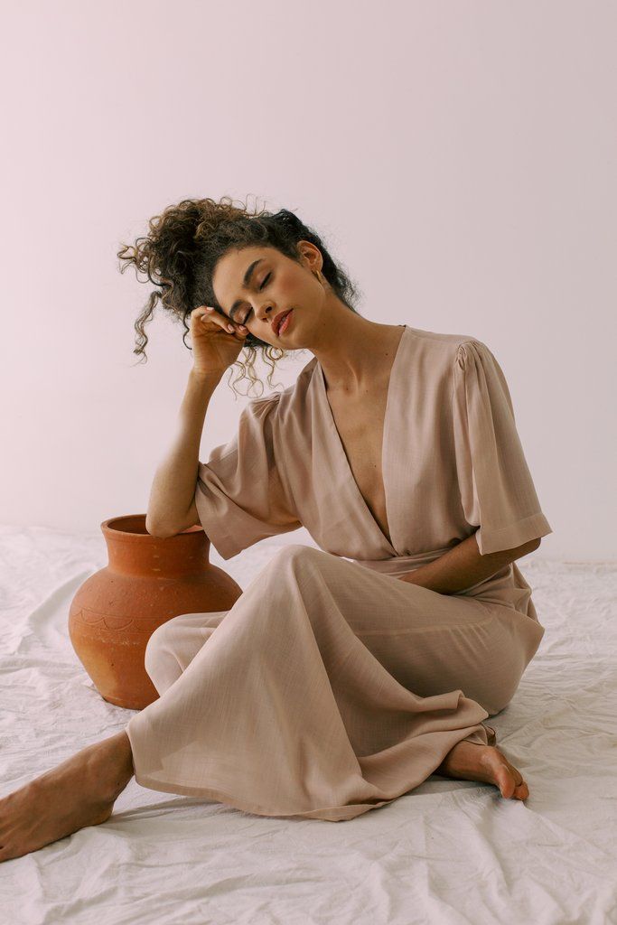 Woman in elegant beige dress sitting, eyes closed, next to clay pot on white sheet. Relaxed and serene atmosphere.