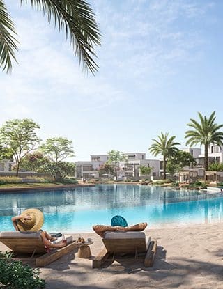 Beachfront view with loungers and palm trees by a clear blue pool, under a sunny sky, near modern villas.