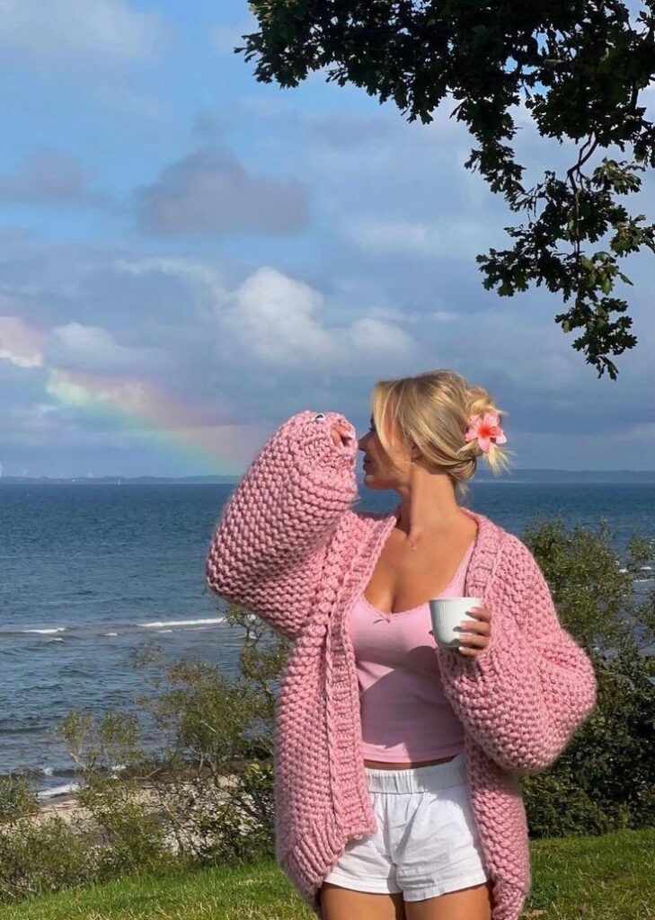 Woman in pink sweater enjoying coffee by ocean, with rainbow in the background.