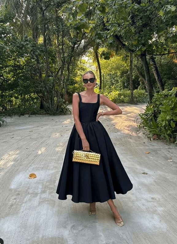 Woman in black dress and sunglasses holding a gold clutch, standing on a tree-lined path in summer.