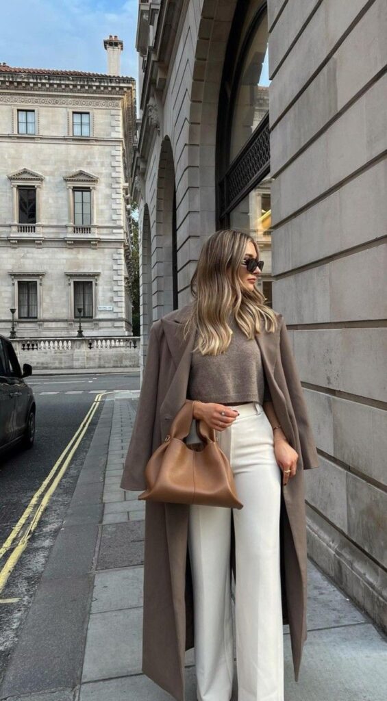 Stylish woman in trench coat and sunglasses, holding a brown tote bag on an urban street with classic architecture.