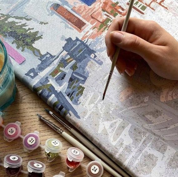 Person painting by numbers with various colorful paints and brushes on a wooden table.