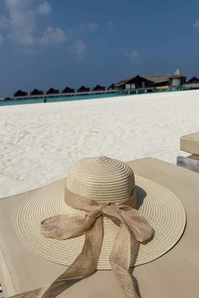Straw hat with ribbon on a sandy beach, overwater bungalows and blue sky in the background.