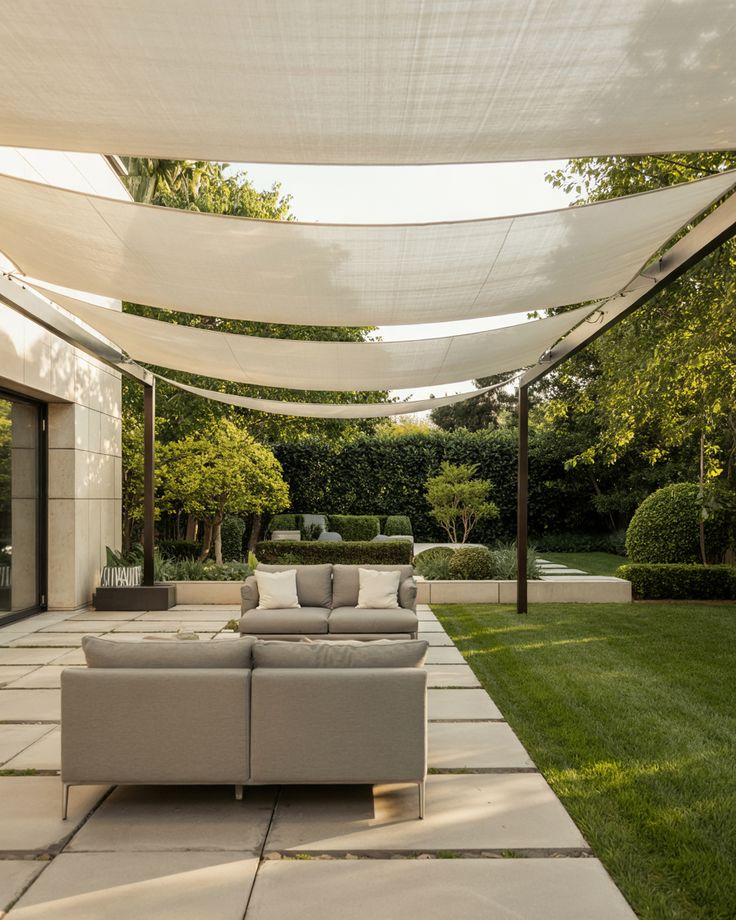 Modern outdoor patio with gray sofas under sunshades, surrounded by green garden and hedges.