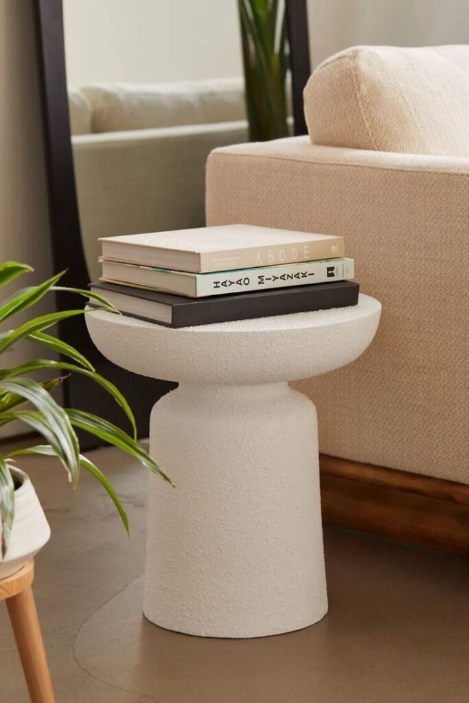 Modern living room with textured white side table and stacked books, next to beige sofa and indoor plants.