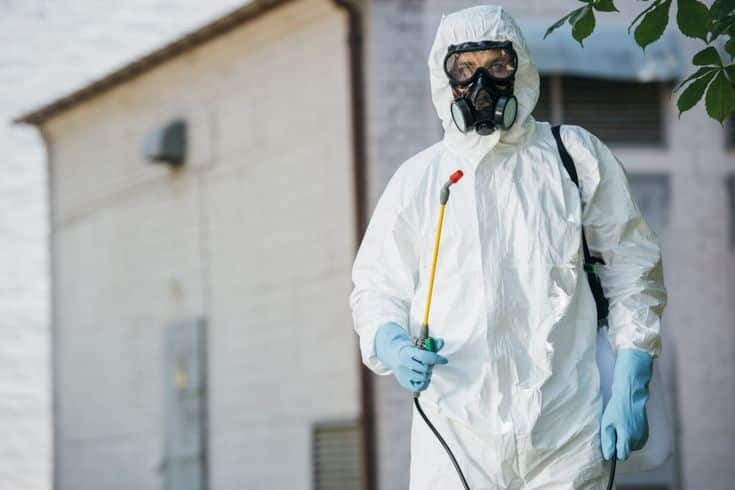 Person in protective gear with spray equipment for pest control near building.