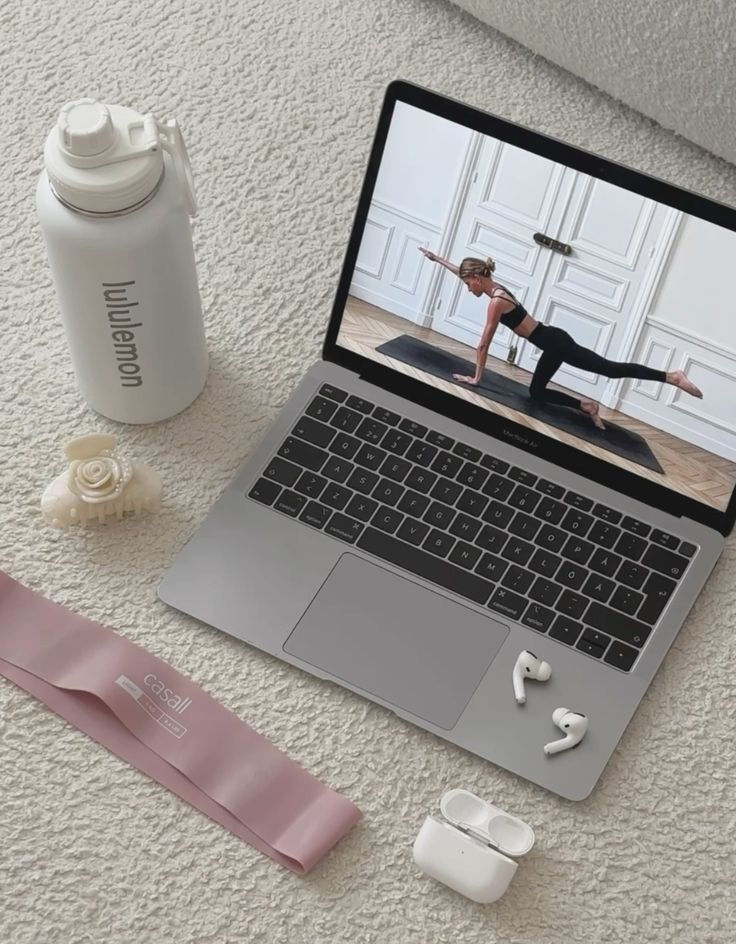 Online workout setup: laptop streaming yoga, water bottle, exercise band, hair tie, and earbuds on a carpet.