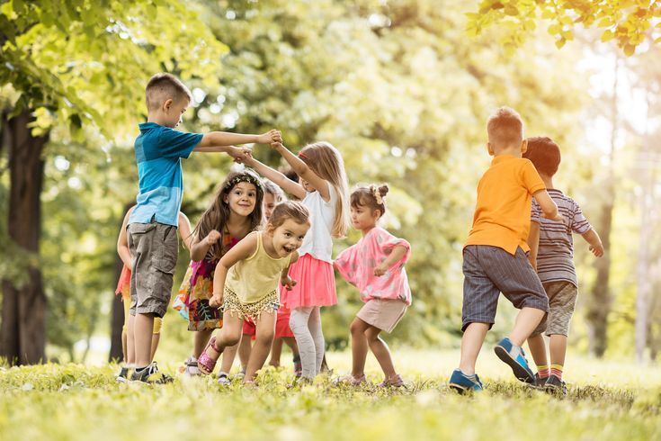 Children playing happily in a circle outdoors on a sunny day, surrounded by trees and greenery.
