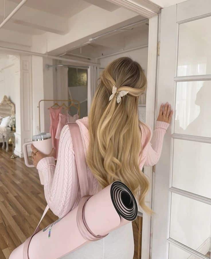 Woman with yoga mat and drink entering a room, styled hair with bow, wearing cozy pink sweater, elegant, serene setting.
