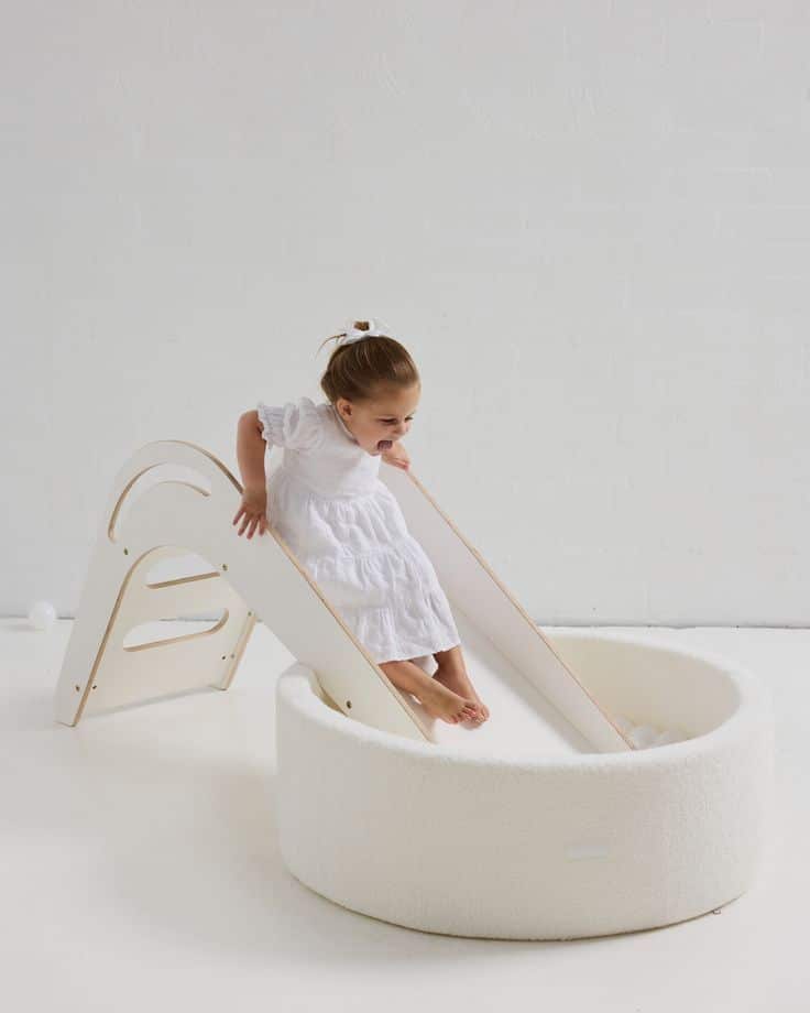 Child playing on a modern white slide with a soft ball pit in a minimalistic room.