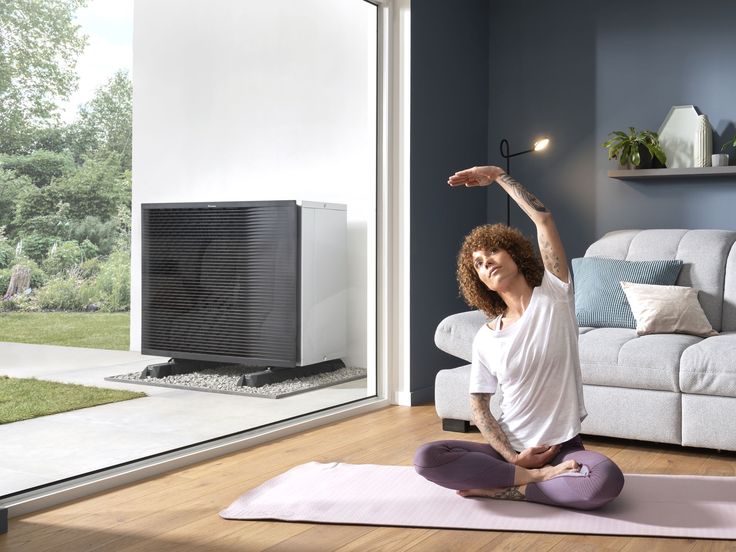 Woman practicing yoga indoors near large window with heat pump outside, modern cozy living space.