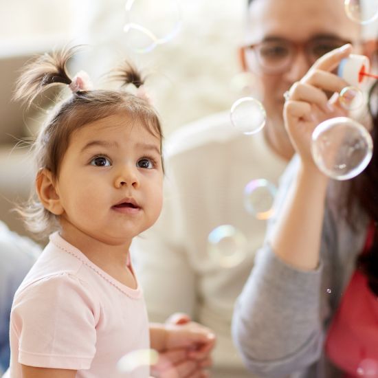 Rainy Day Indoor Play Guide Toddler with pigtails watching bubbles indoors with family, wearing a pink shirt and looking curious. | Sky Rye Design Toddler with pigtails watching bubbles indoors with family, wearing a pink shirt and looking curious.