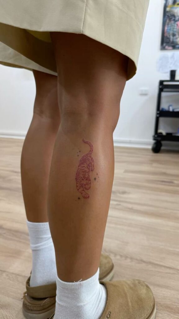 Minimalistic pink tiger tattoo on calf with white socks and beige shoes, hardwood floor background.
