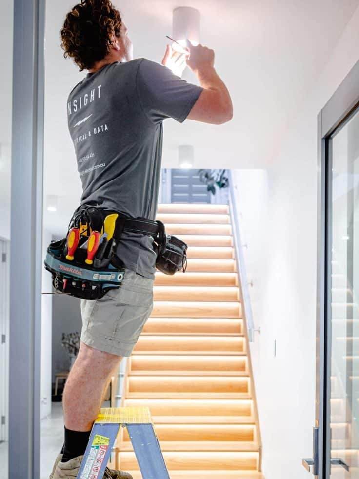 Electrician on ladder fixing light fixture in modern hallway, tool belt visible.