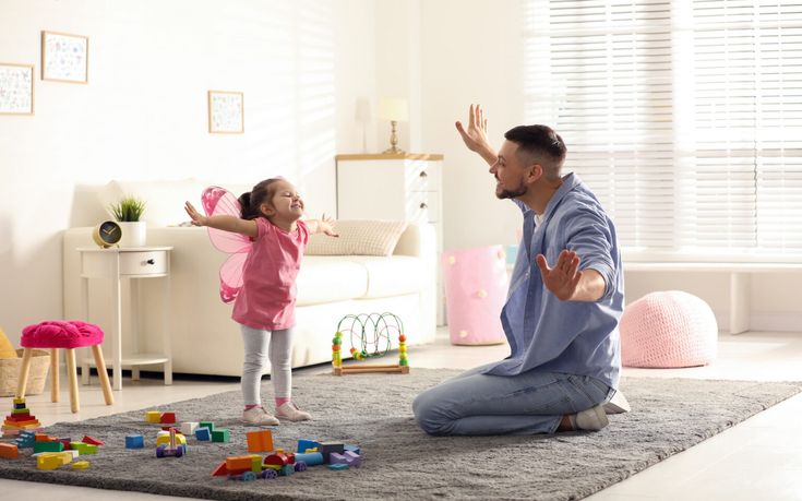 Father and daughter playing joyfully in a bright living room with toys scattered on the floor.