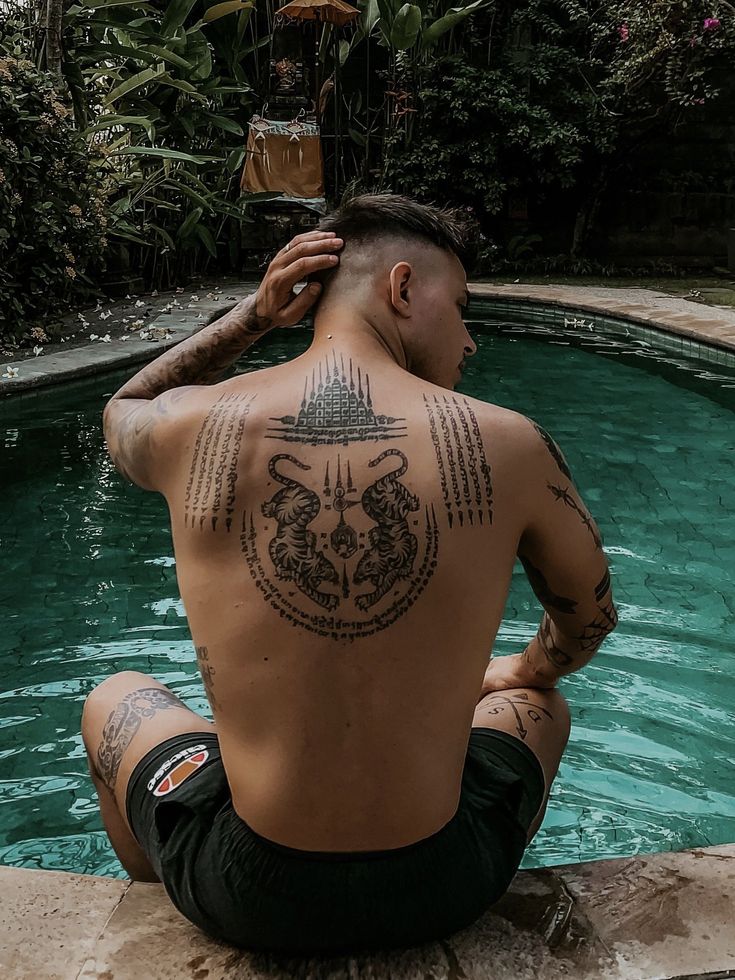 Man with intricate back tattoos sits by a serene pool surrounded by lush greenery.