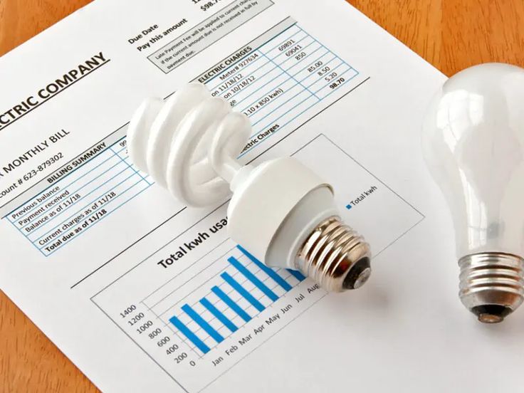 Electric bill with bar graph, CFL bulb, and incandescent bulb on a wooden table, showcasing energy usage comparison.