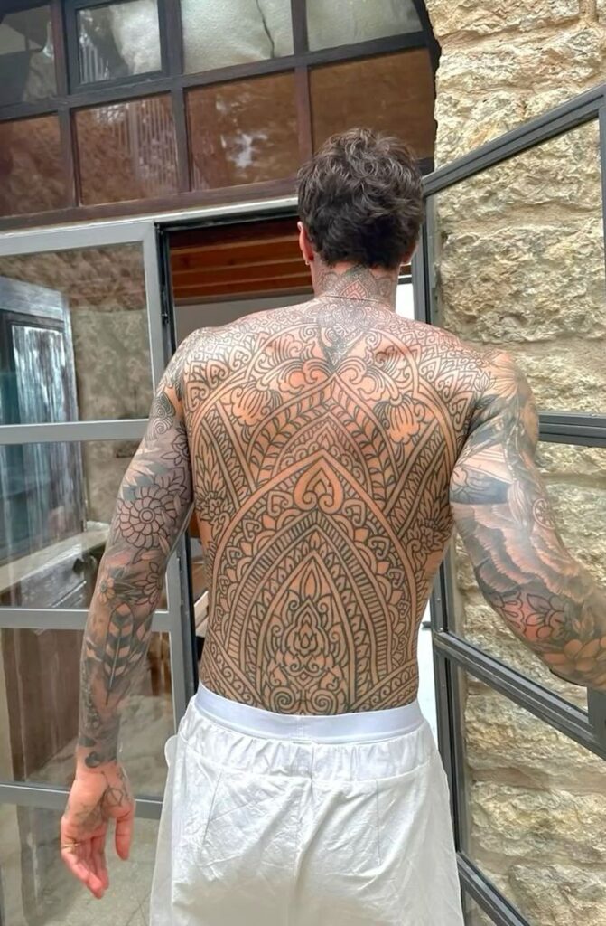 Man with intricate full-back tattoo art, standing by a glass door in a stone-walled room.