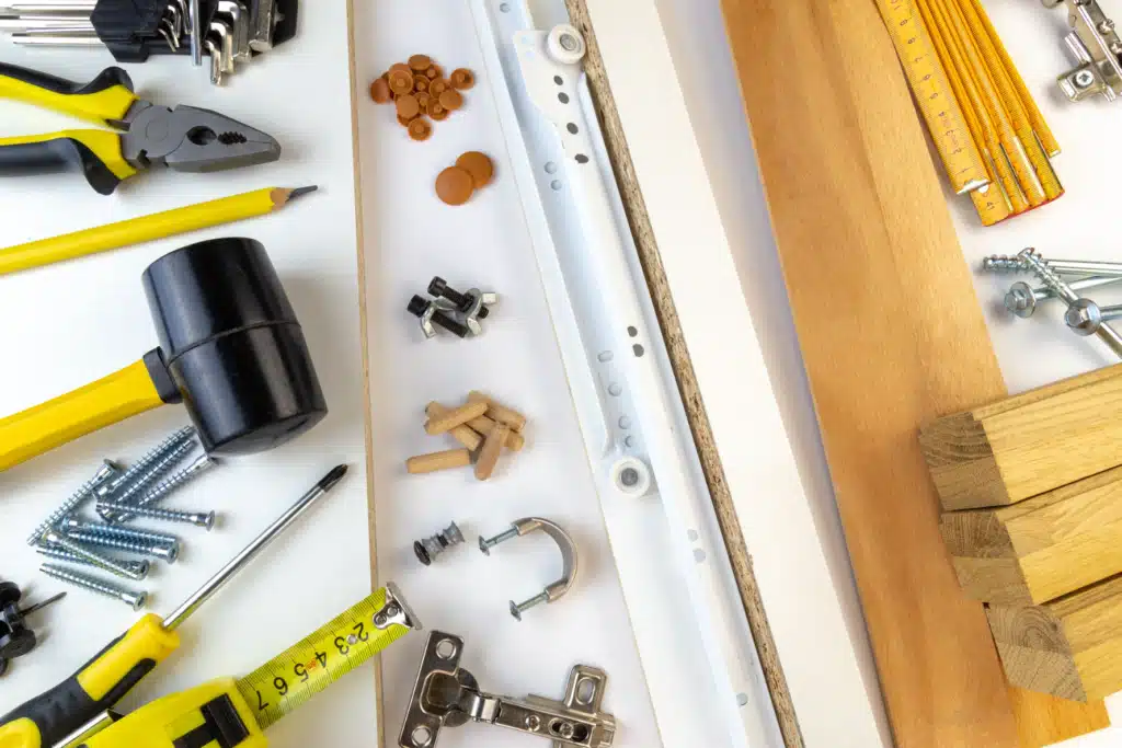Assorted DIY tools and hardware on white background, including pliers, screws, wooden pieces, and a tape measure.