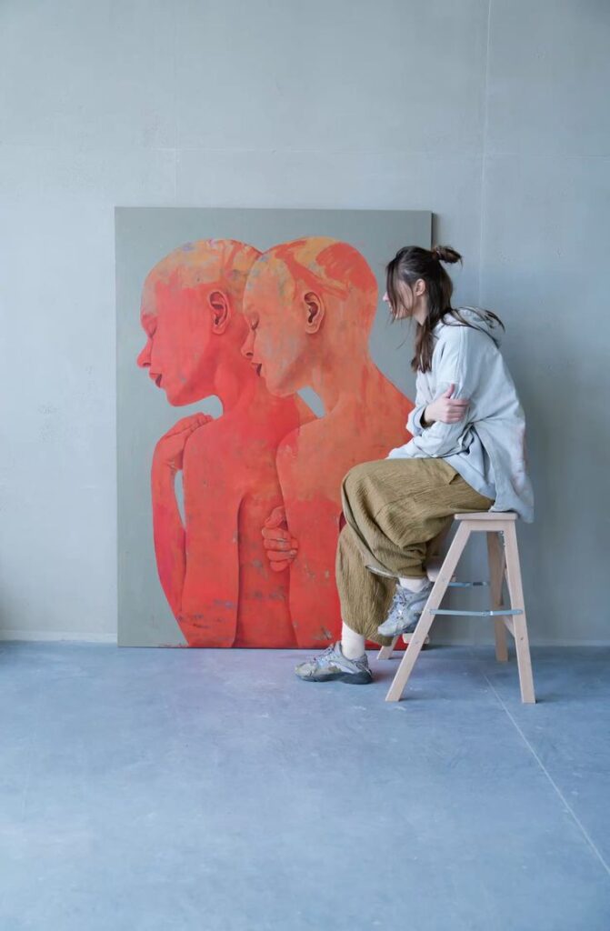 Artist contemplating a vibrant, red-toned dual portrait painting while seated on a wooden stool in a minimalist studio. Drawing aesthetics