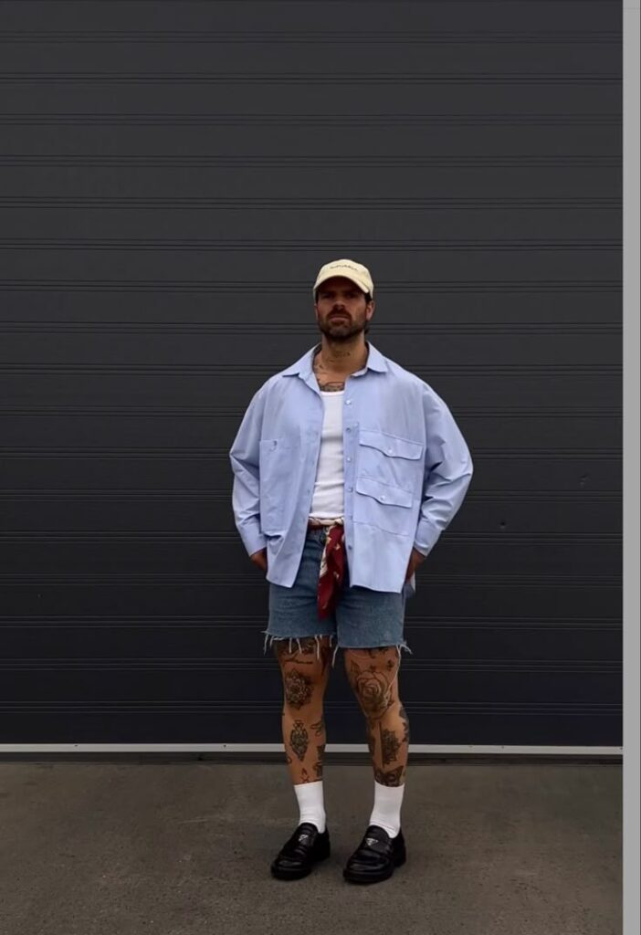 Man in casual outfit with tattoos, wearing a cap, blue shirt, denim shorts, and loafers, stands against gray background.