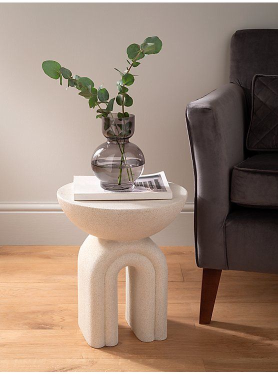 Modern side table with a glass vase and eucalyptus leaves, next to a gray armchair, on wooden floor in cozy room.